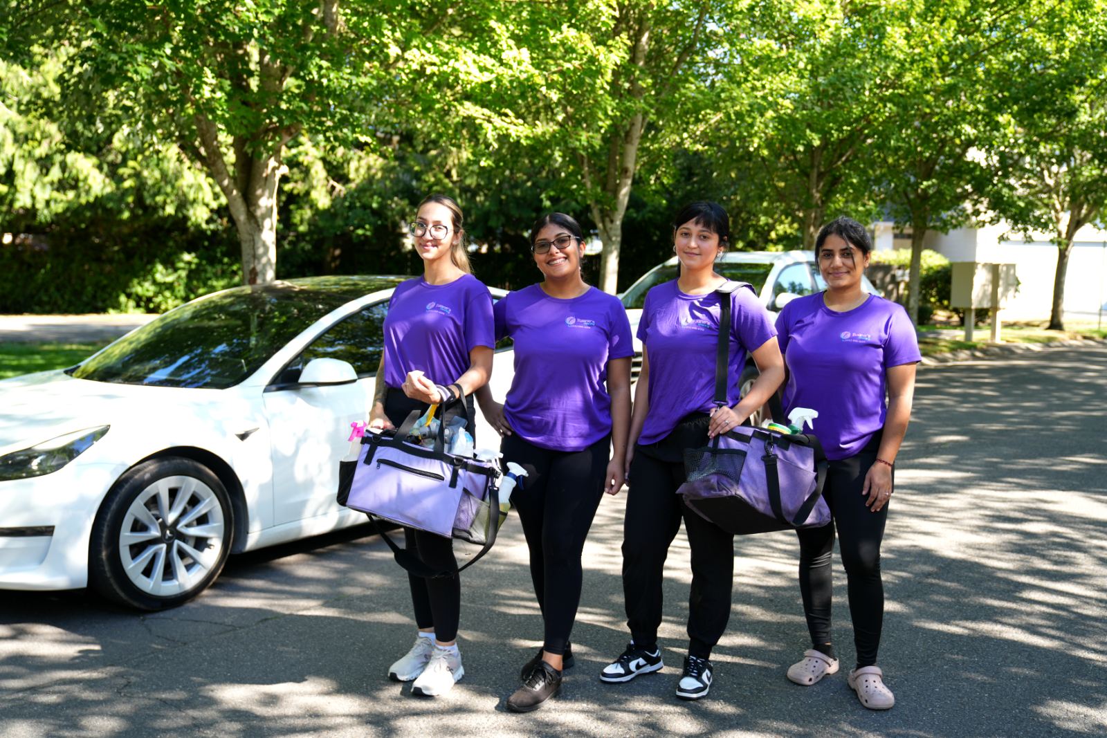 Four Tuane's Cleaning team members in purple shirts on a residential street, holding their supply bags.